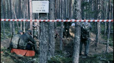 Skupina mužů v maskovacím oblečení v lese, prostor ohraničen výstražnou páskou.