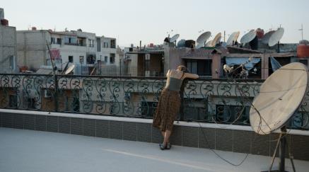 Woman leaning on balcony railing overlooking cityscape with satellite dishes.