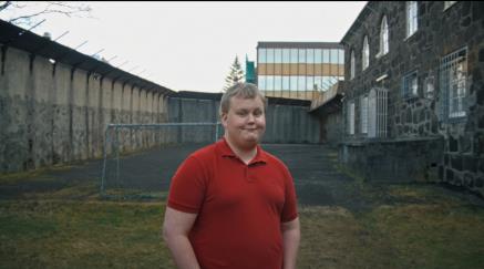 Muž v červeném tričku stojí před budovou s velkými okny za šera.
