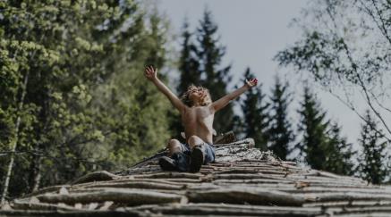 Chlapec s radostě zdviženýma rukama sedí na hromadě dříví v lese.