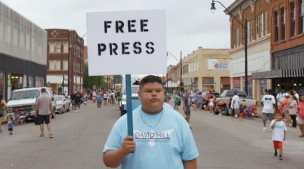 Chlapec drží ceduli s nápisem "FREE PRESS" na ulici plné lidí.
