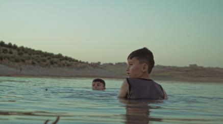 Dva chlapci plavou ve vodě, jeden z nich se dívá přes rameno.
