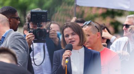 Woman speaking into a microphone at an outdoor event with a camera nearby.